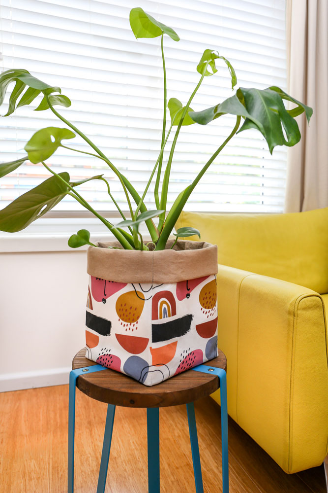 monstera-plant-in-terracotta-muted-rainbow-plant-pot-cover-placed-on-a-wooden-stool-next-to-a-yellow-sofa