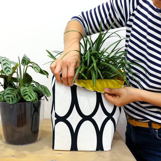 woman-folding-the-top-of-a-white-black-and-yellow-fabric-planter-featuring-geometric-circles-fabric-pattern-with-two-potted-plants-on-a-table